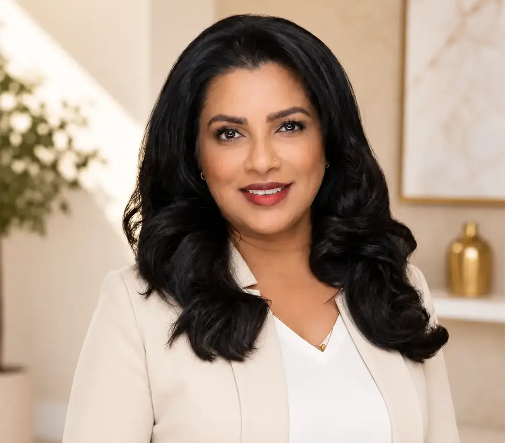 Professional portrait of Nicola Aulds, San Diego real estate agent, wearing a cream blazer with long, dark wavy hair against an elegant, softly blurred interior background.